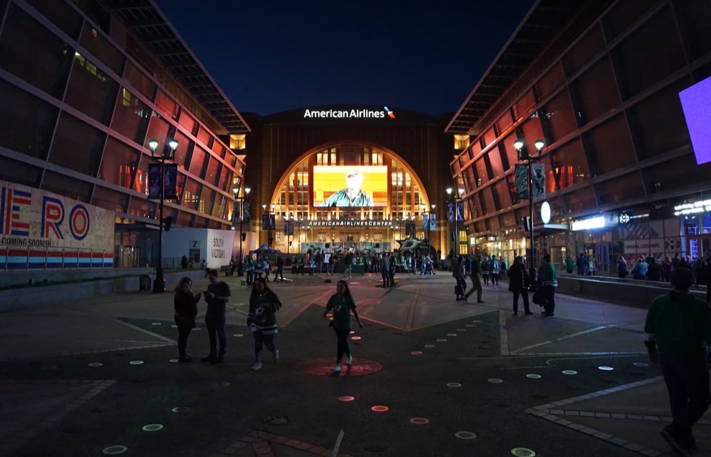 Fans in Dallas Stars jerseys cross the plaza in front of the lit entrance arch of American Airlines Center at night