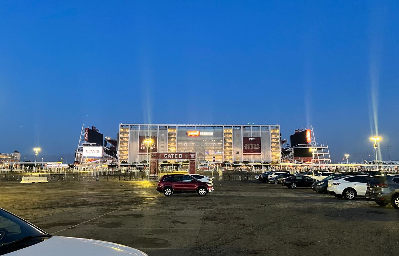 Levi’s Stadium at dusk seen across a parking lot, with the Gate B arch and a lit 49ers banner on its glass facade