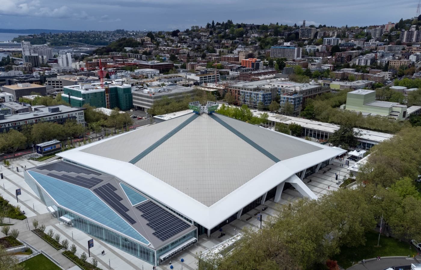 Aerial view of Climate Pledge Arena’s roof and glass-walled entrance on the Seattle Center campus, with Queen Anne Hill behind it