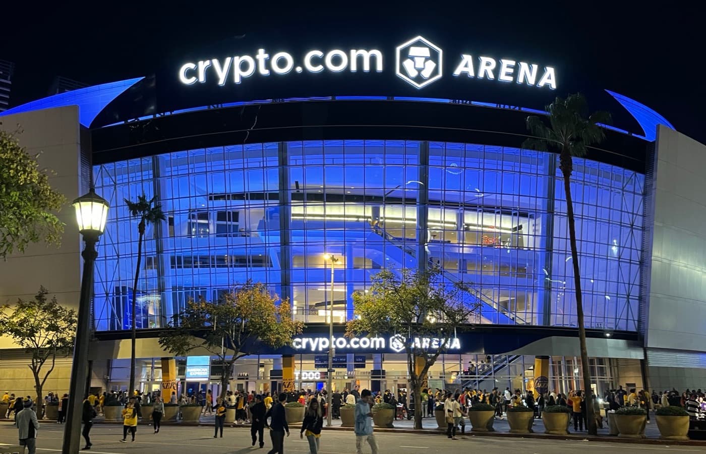Crypto.com Arena’s illuminated entrance and signage at night, with palm trees and a crowd of fans outside.