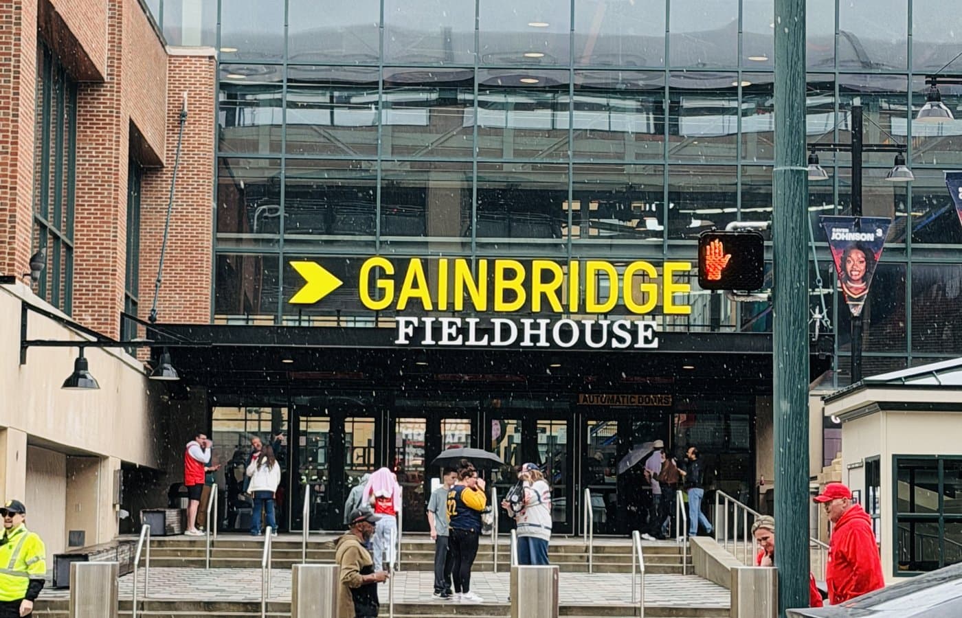 The Gainbridge Fieldhouse entrance plaza in downtown Indianapolis, with the arena’s yellow-and-white sign above the doors and fans gathering outside in light rain