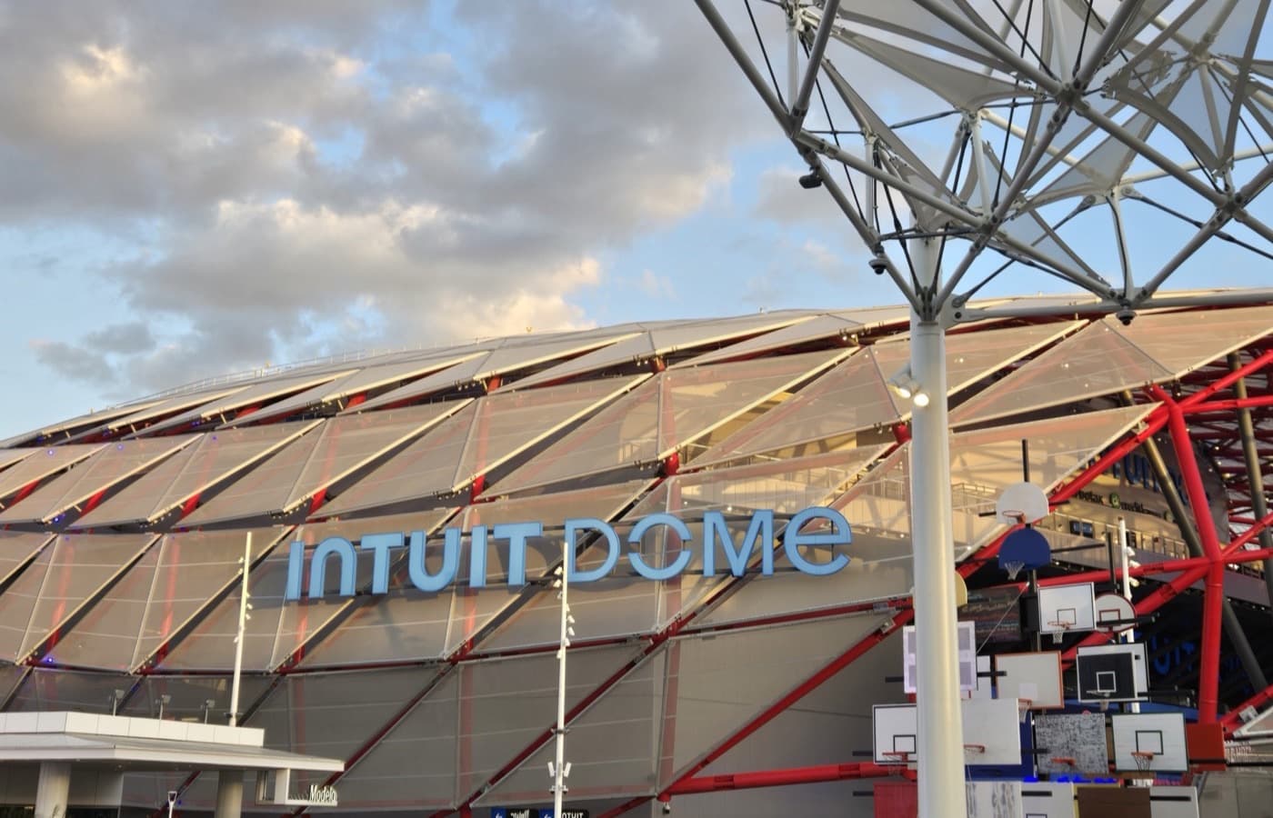 Intuit Dome’s angular exterior and illuminated sign, seen from the arena plaza with a sculptural basketball-hoop installation in the foreground.