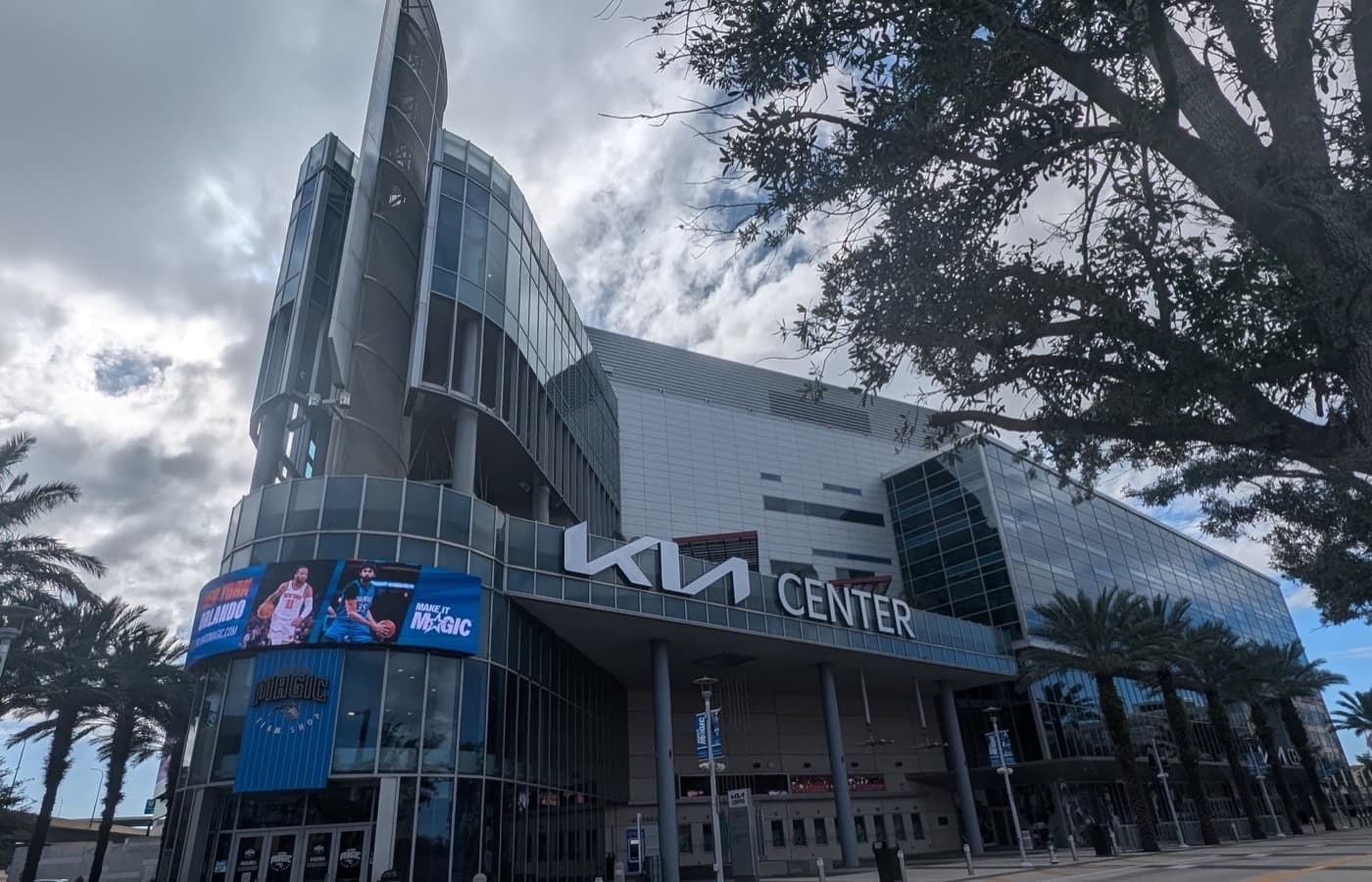Exterior of Kia Center in downtown Orlando, with the arena’s Kia sign over the glass entrance and palm trees along the street