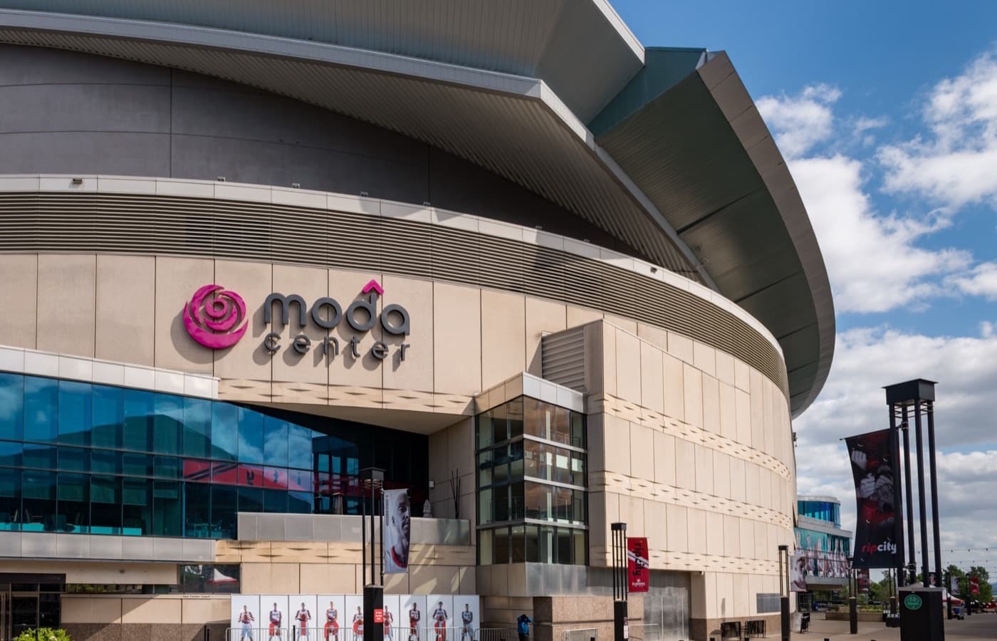The curved tan exterior of Moda Center in Portland, with its rose logo and Rip City banners over the main entrance, under a blue sky