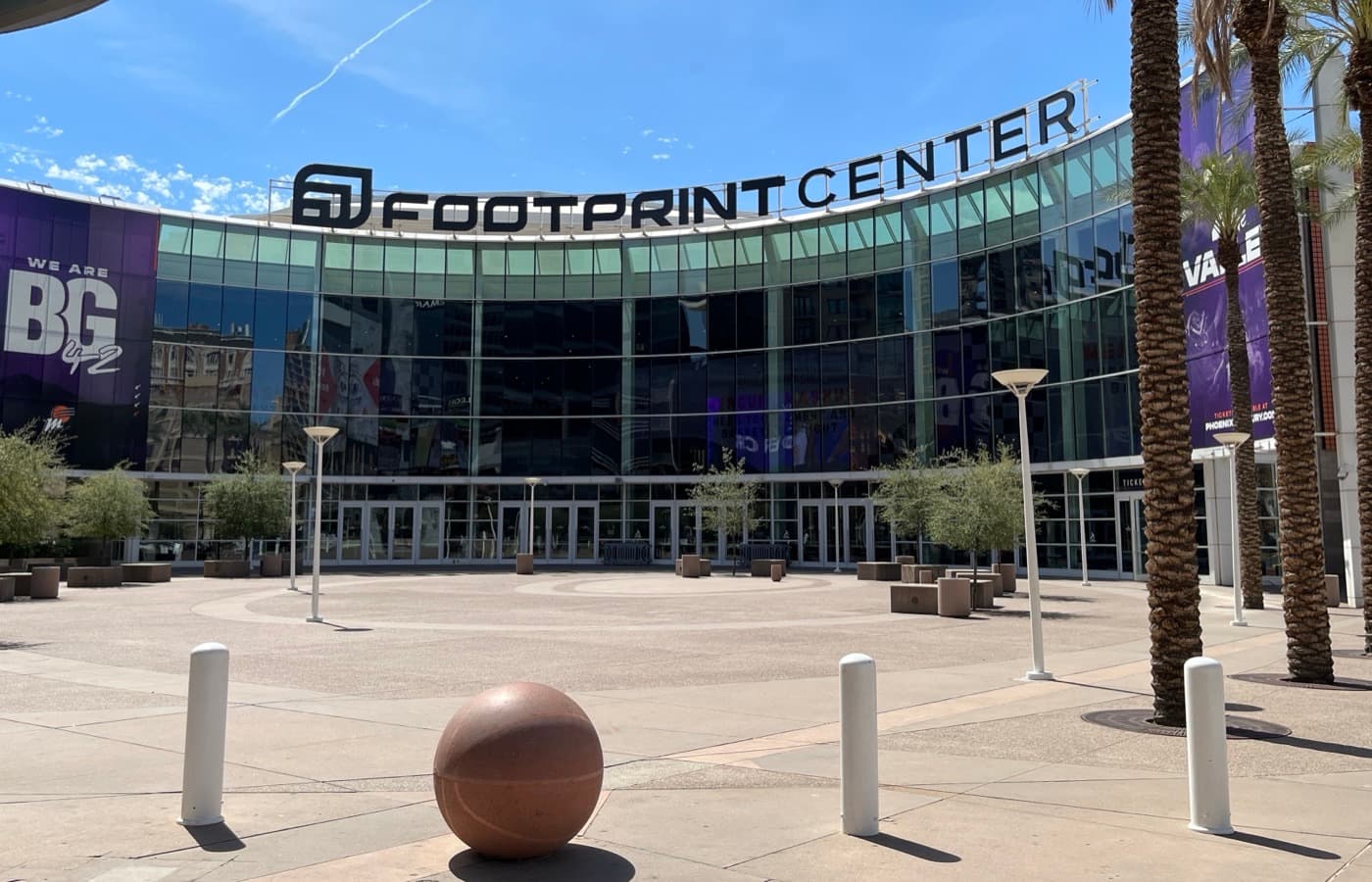 The arena’s glass-fronted main entrance plaza in downtown Phoenix, under its former Footprint Center signage, with palm trees, lamp posts and a basketball sculpture in the foreground