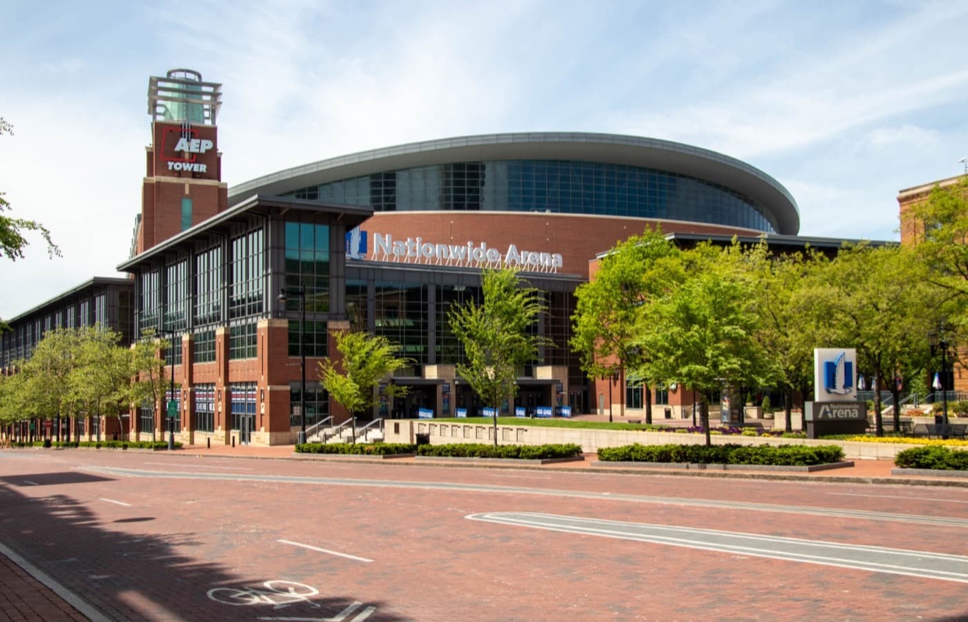 Nationwide Arena’s brick-and-glass entrance and marquee in Columbus’s Arena District, with the AEP Tower on the left and trees along a brick-paved street