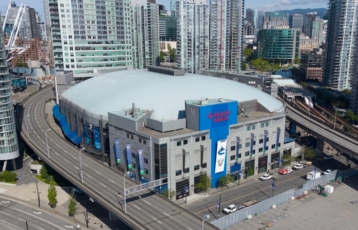 Aerial view of Rogers Arena and its white roof among downtown Vancouver high-rises, with an elevated roadway curving past the building