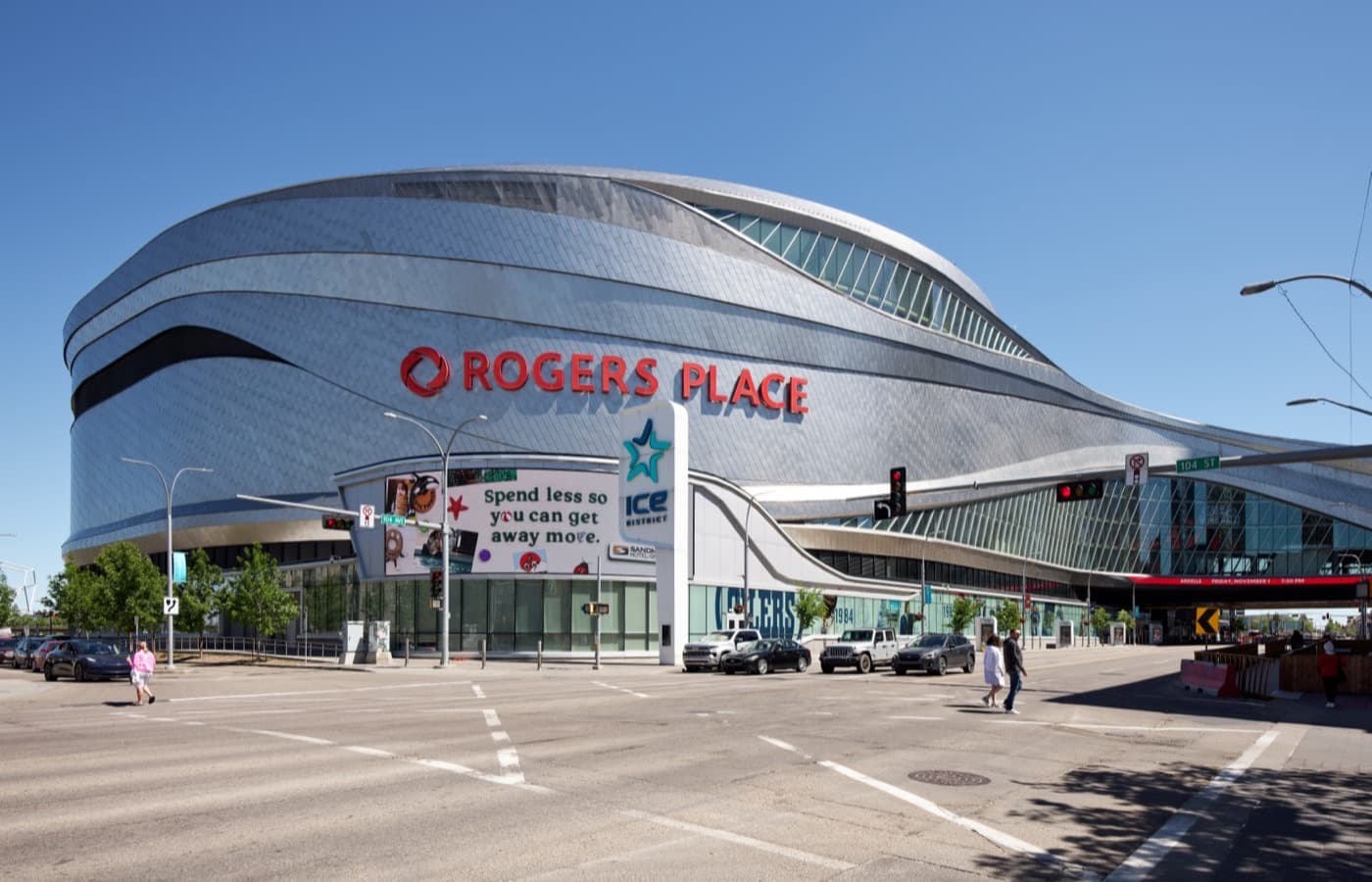 The curved metal-clad exterior of Rogers Place in downtown Edmonton, with the red Rogers Place sign and an ICE District sign at street level on a clear day