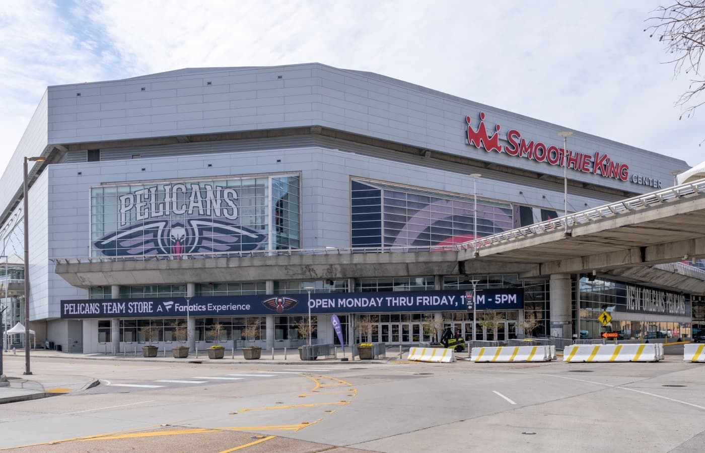 The Smoothie King Center’s metal-panel exterior in New Orleans, with the arena’s sign and a New Orleans Pelicans logo across the upper windows above the team store entrance