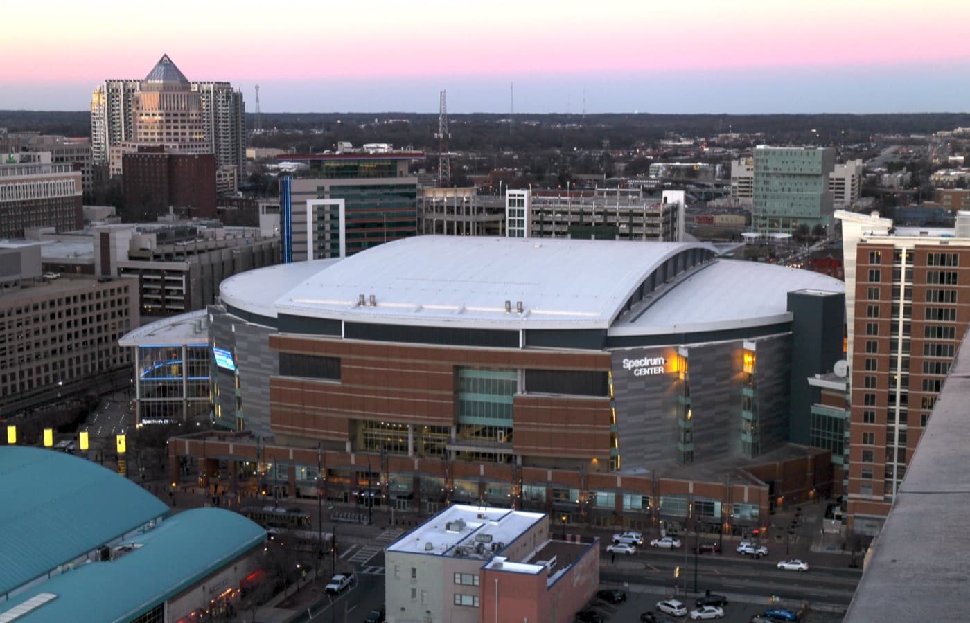 Spectrum Center at dusk, seen from a rooftop in Uptown Charlotte, with its curved white roof, lit sign and surrounding high-rises