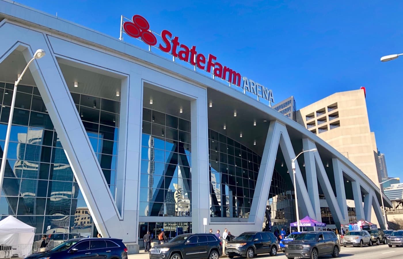 State Farm Arena’s angular glass-and-steel entrance in downtown Atlanta, with the State Farm sign above the doors and cars parked along the street outside