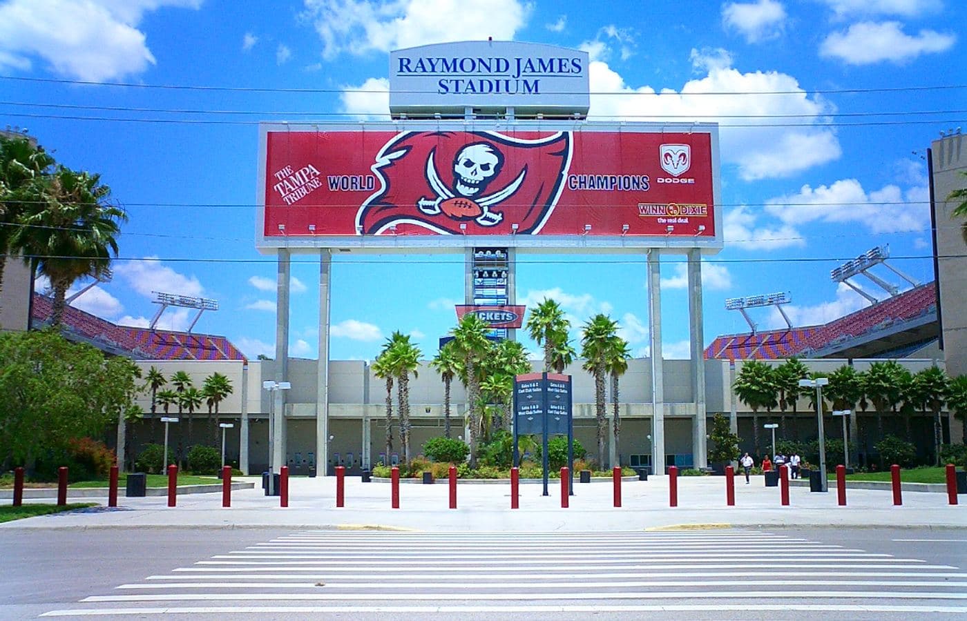 The Raymond James Stadium sign and a Buccaneers “World Champions” banner above the palm-lined plaza at the stadium entrance, seen from across the street