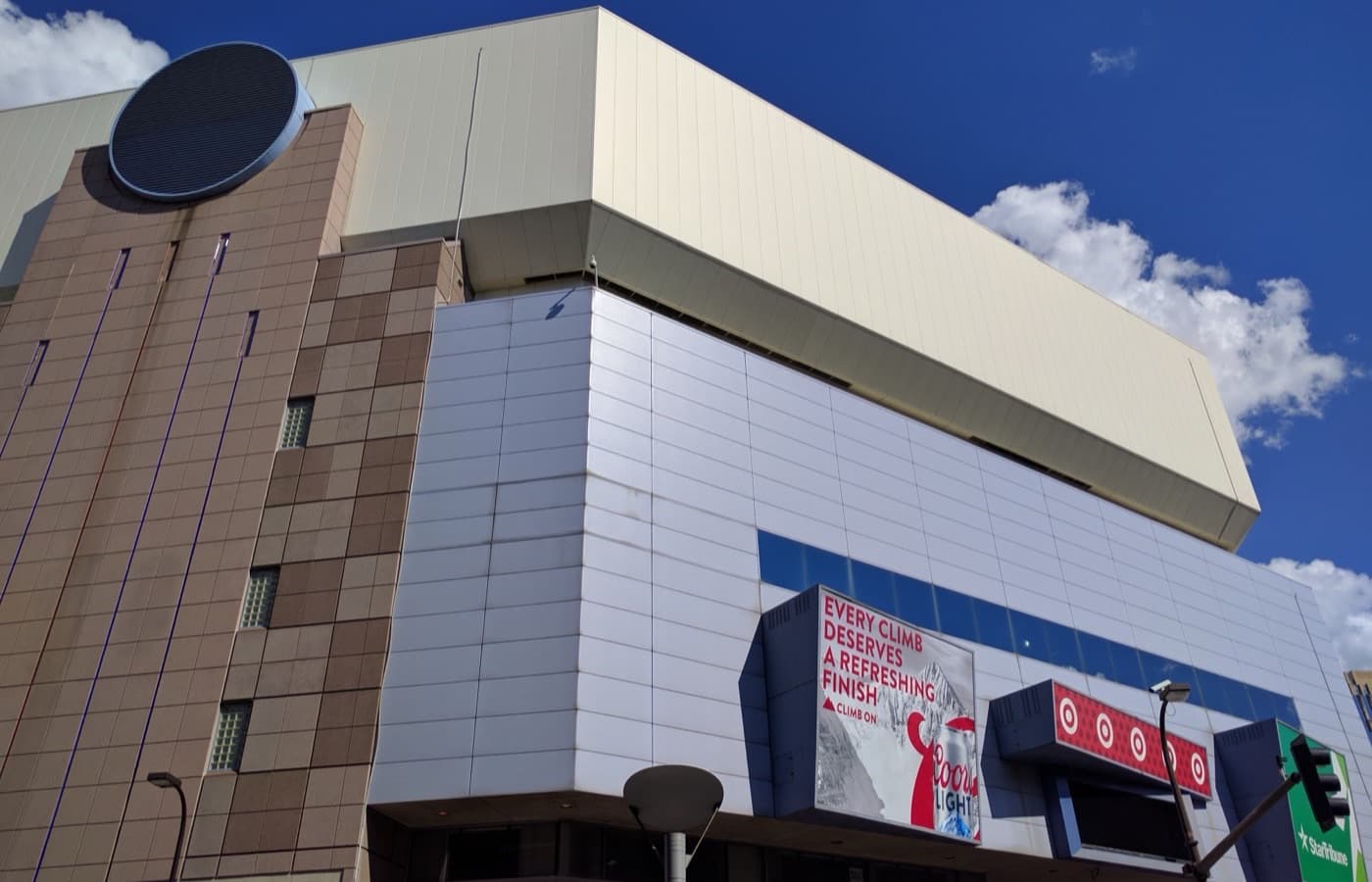 A low-angle view of Target Center’s exterior in downtown Minneapolis, with its paneled upper level and a lit Target sign above the street-level entrance, against a blue sky